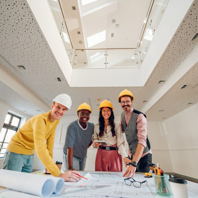 Multiracial team of architects having a meeting while using blue Multiracial group of architects gathering at table with papers and gadgets while sharing ideas about new project they are working on. Looking into the camera. Modern building design. Copy space.